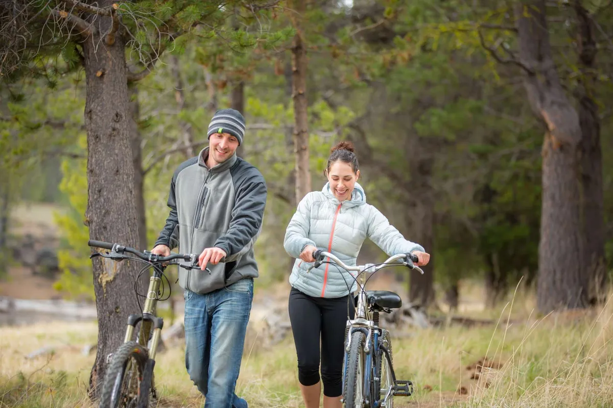 Two people walk with bicycles along a grassy path under tall trees, enjoying a joyful and relaxed moment exploring nature together.