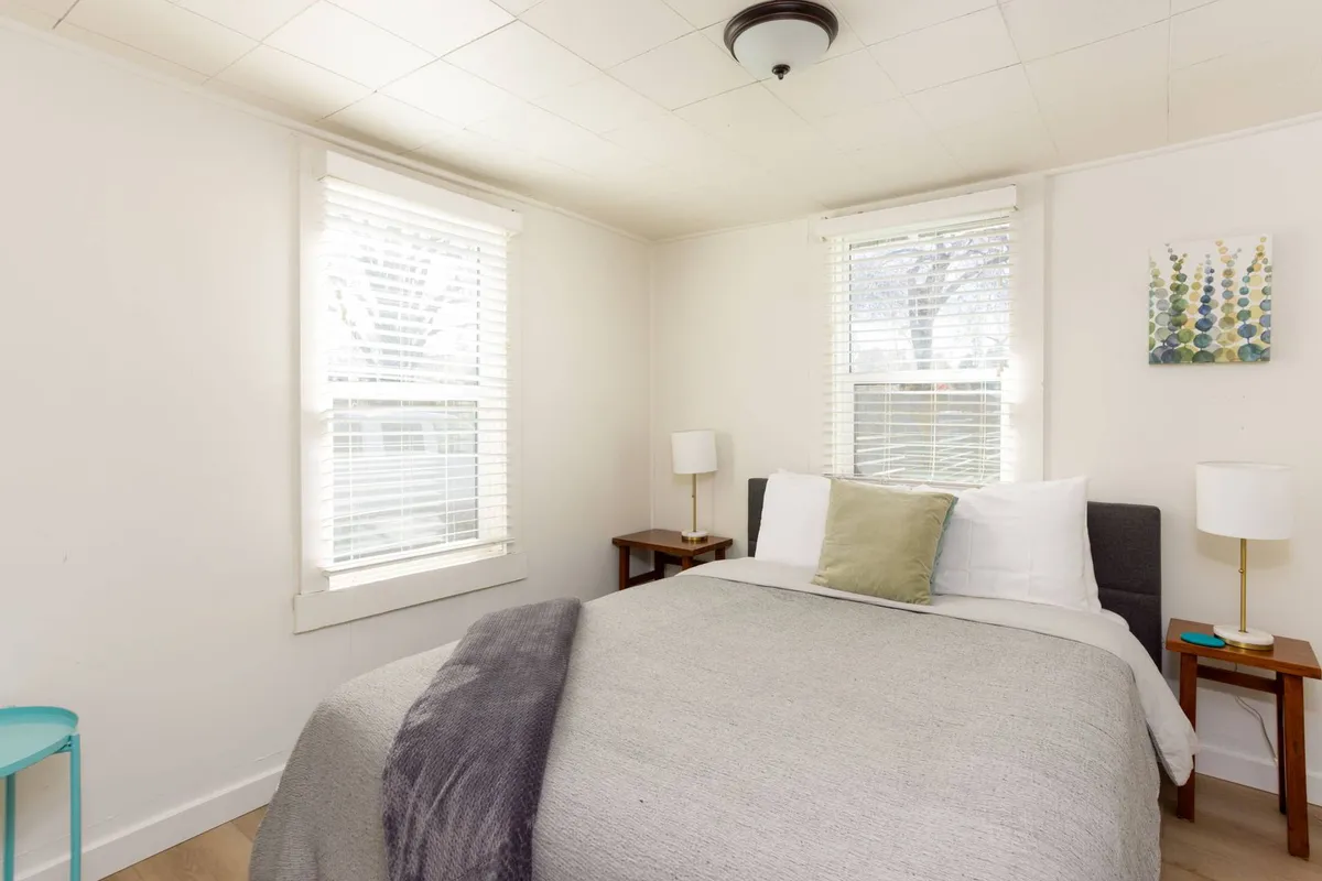 A serene bedroom featuring a queen-size bed adorned with a gray blanket and plush pillows. Natural light pours in from two windows, illuminating the space and highlighting colorful artwork above the bed. Nightstands with lamps and a turquoise side table add comfort and character.