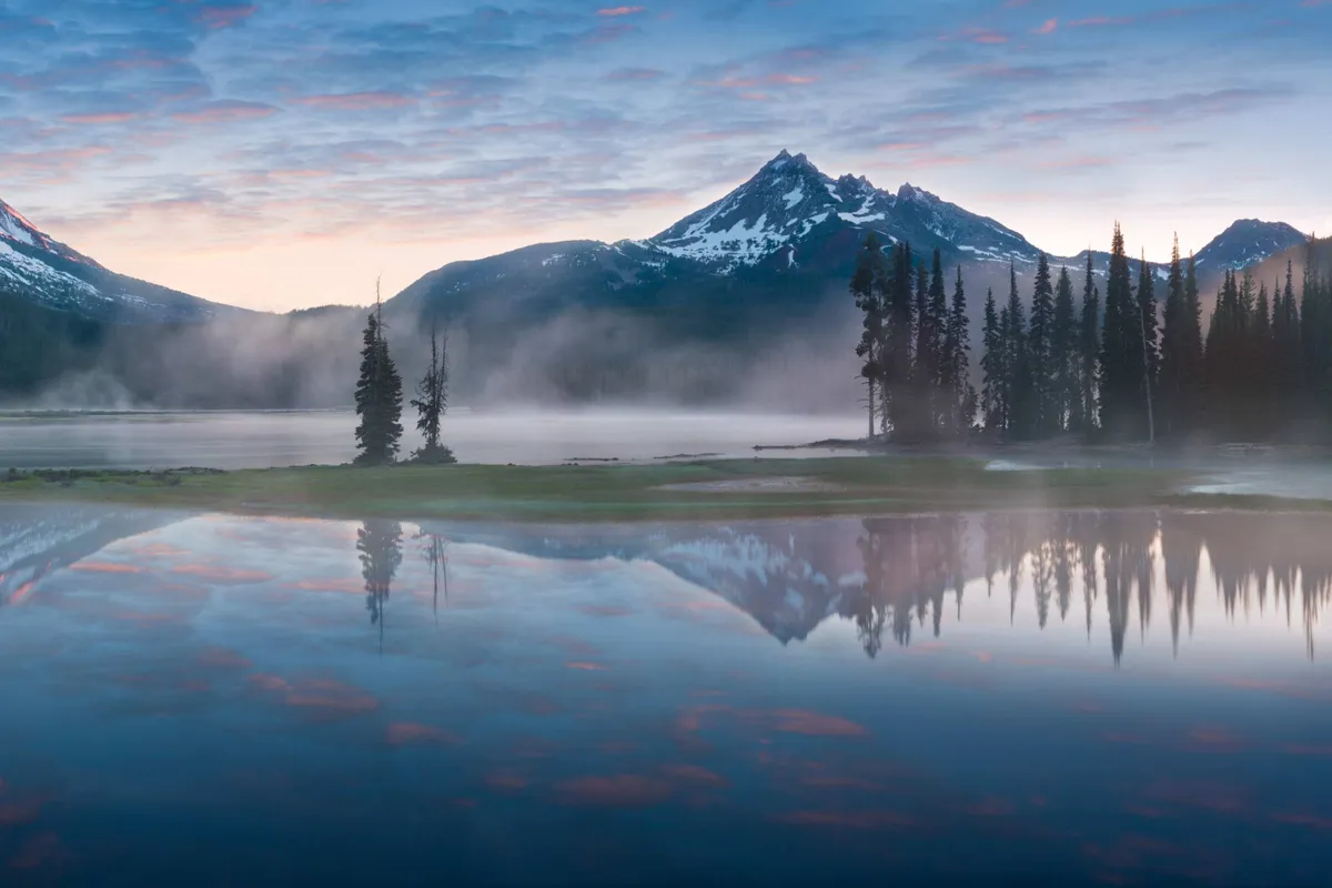 A sweeping view of tranquil mountain peaks reflecting in a pristine lake, bordered by evergreen trees and an open sky. The scene evokes the peacefulness of the Cascade Lakes and high mountain environments, inviting exploration and relaxation surrounded by natural beauty.
