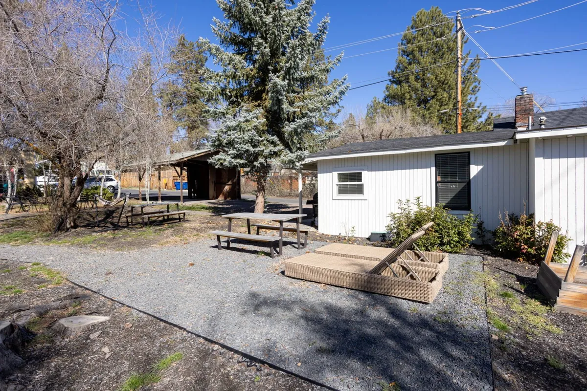 Set in the backyard, this image shows picnic tables, a gravel pathway, lounge chairs, and mature trees, creating a casual, inviting environment for outdoor dining, relaxation, and social gatherings. The backyard is bathed in natural light, further enhancing its appeal for after-work unwinding or weekend leisure.