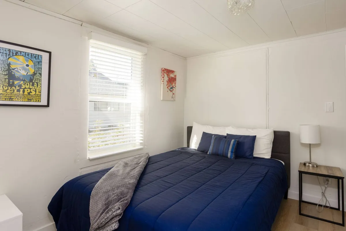 An inviting bedroom with a queen-size bed dressed in blue bedding, complemented by framed artwork and a nightstand with a lamp. Soft morning light filters through the window, emphasizing the space’s relaxed and welcoming mood.