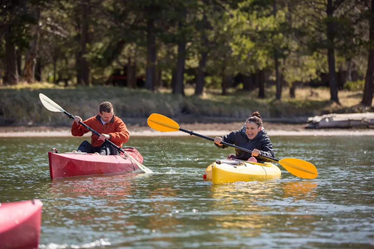 Two kayakers paddle across calm water, surrounded by lush green trees. The tranquil setting captures the joy of outdoor water activities and the beauty of the Deschutes River environment. The scene conveys a sense of adventure and connection to nature, ideal for those seeking memorable experiences just minutes from the home.