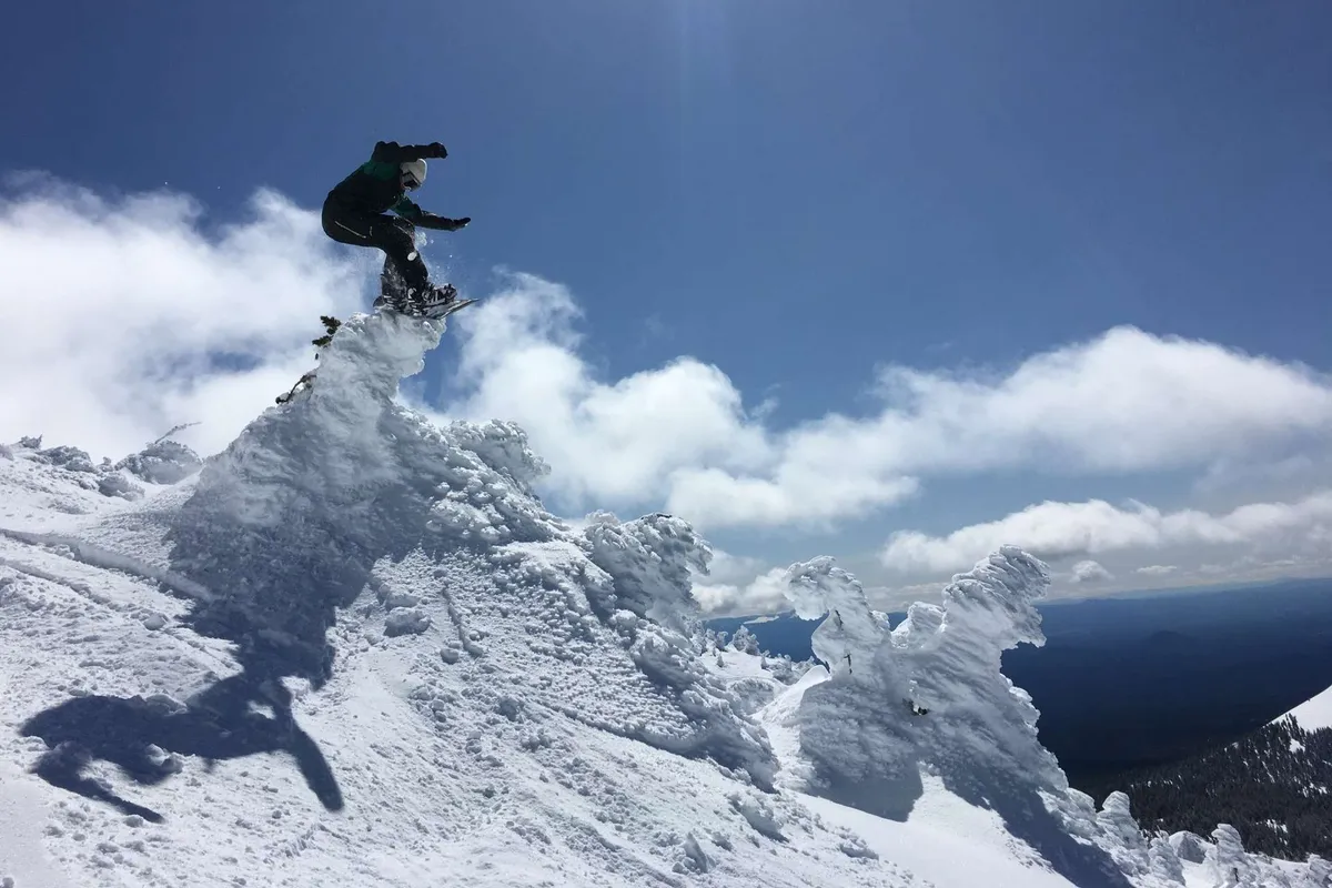 An exhilarating view of a snowboarder mid-jump on a snow-covered mountain peak, set against a dramatic sky with clouds. The expansive snowfields and visible camaraderie among snow enthusiasts capture the spirit of winter adventure in Bend.