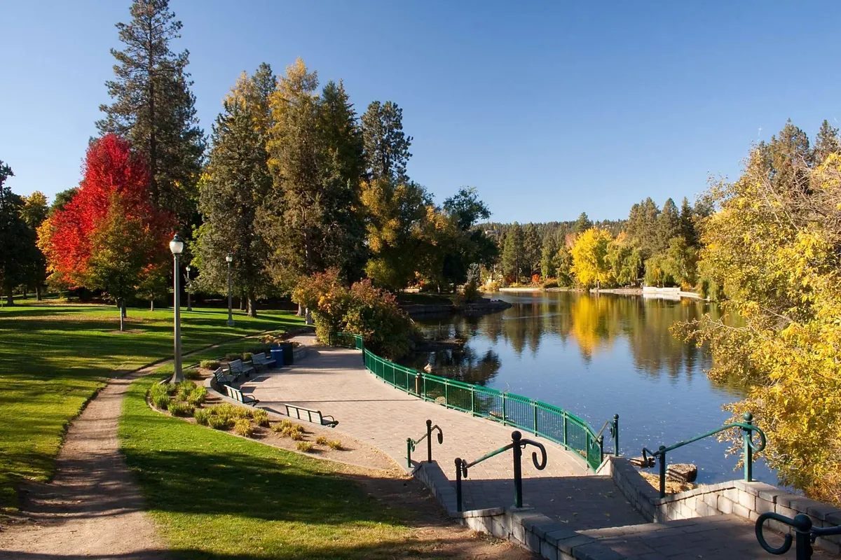 A tranquil outdoor scene featuring a winding pathway beside a calm water surface, bordered by vibrant autumn trees and several park benches. The area is ideal for walking, jogging, or quietly taking in the natural scenery, with park-like features that evoke opportunities for outdoor adventure.