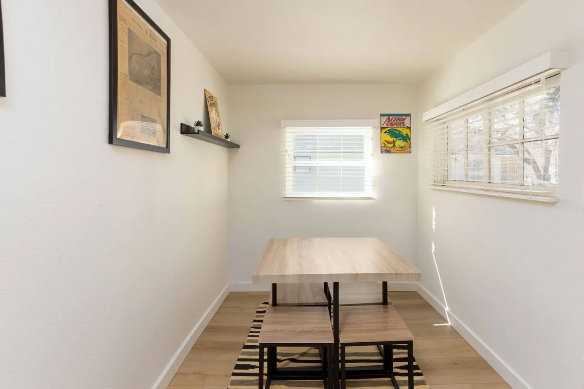 A multipurpose interior with a wooden table, stacked chairs, framed artwork, and a shelf with decor—an adaptable space perfect for casual dining, quiet reading, or working from home.