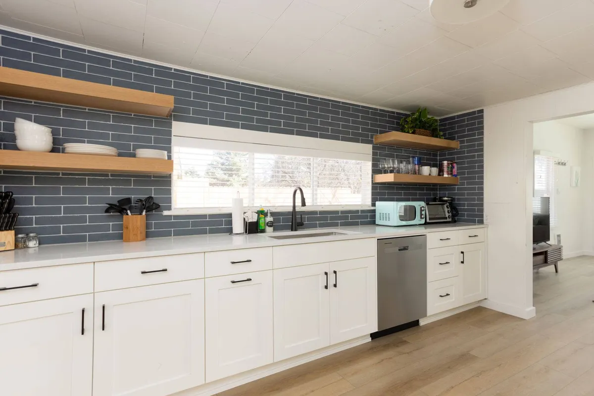 This interior image features a kitchen sink set against a blue tiled backsplash, open shelving, white cabinetry, and stainless steel appliances. The scene is warm and modern, showcasing an inviting, lived-in kitchen ready for meal preparation and gatherings.