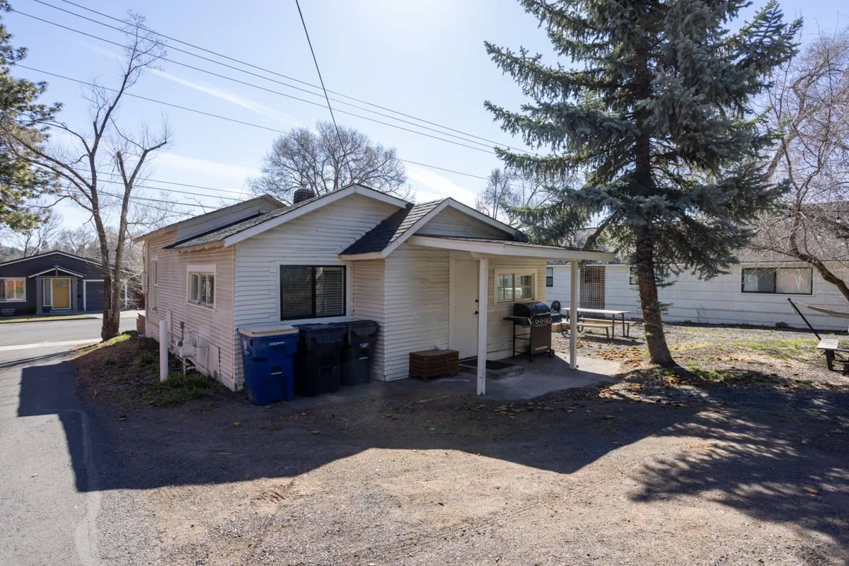 A welcoming exterior view of the house, surrounded by trees and featuring a backyard area with a grill and space for outdoor activities. The friendly, homey atmosphere hints at a private, fenced yard suitable for dogs to play or families to relax together around the firepit.