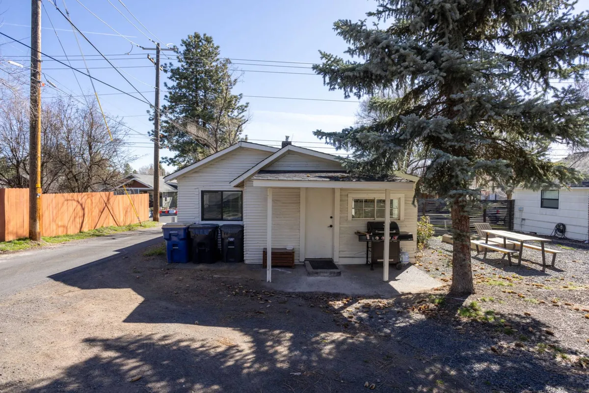 This image captures the welcoming exterior of the property, featuring a sunny front porch with comfortable seating, a picnic table perfect for outdoor meals, and recycling bins indicating a lived-in, community-oriented atmosphere. The scene suggests accessible outdoor spaces ideal for both work breaks and casual gatherings.