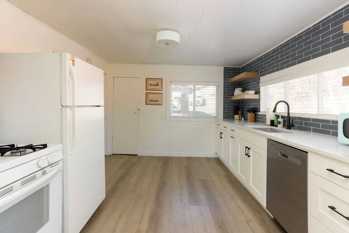 A bright, welcoming kitchen interior with white cabinetry, blue tiled backsplash, stainless steel appliances, and open shelving. The layout is functional, ideal for meal prep, and fits the 'eat-in' concept. The scene feels ready for social cooking or casual dining moments.