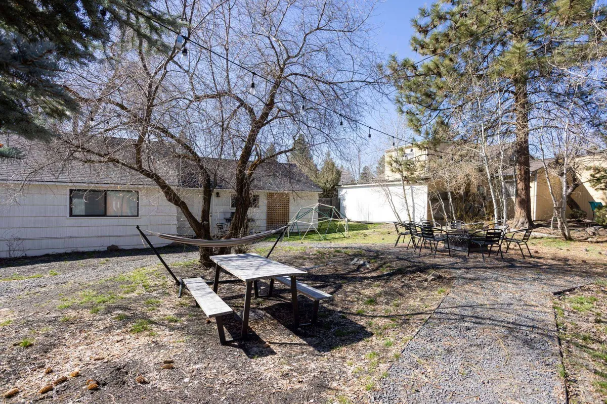 A welcoming backyard scene featuring a hammock, picnic table, string lights, patio seating, and trees. The area is set up for relaxation and social gatherings, inviting guests to sit under the evening lights, dine outdoors, or unwind in the hammock.