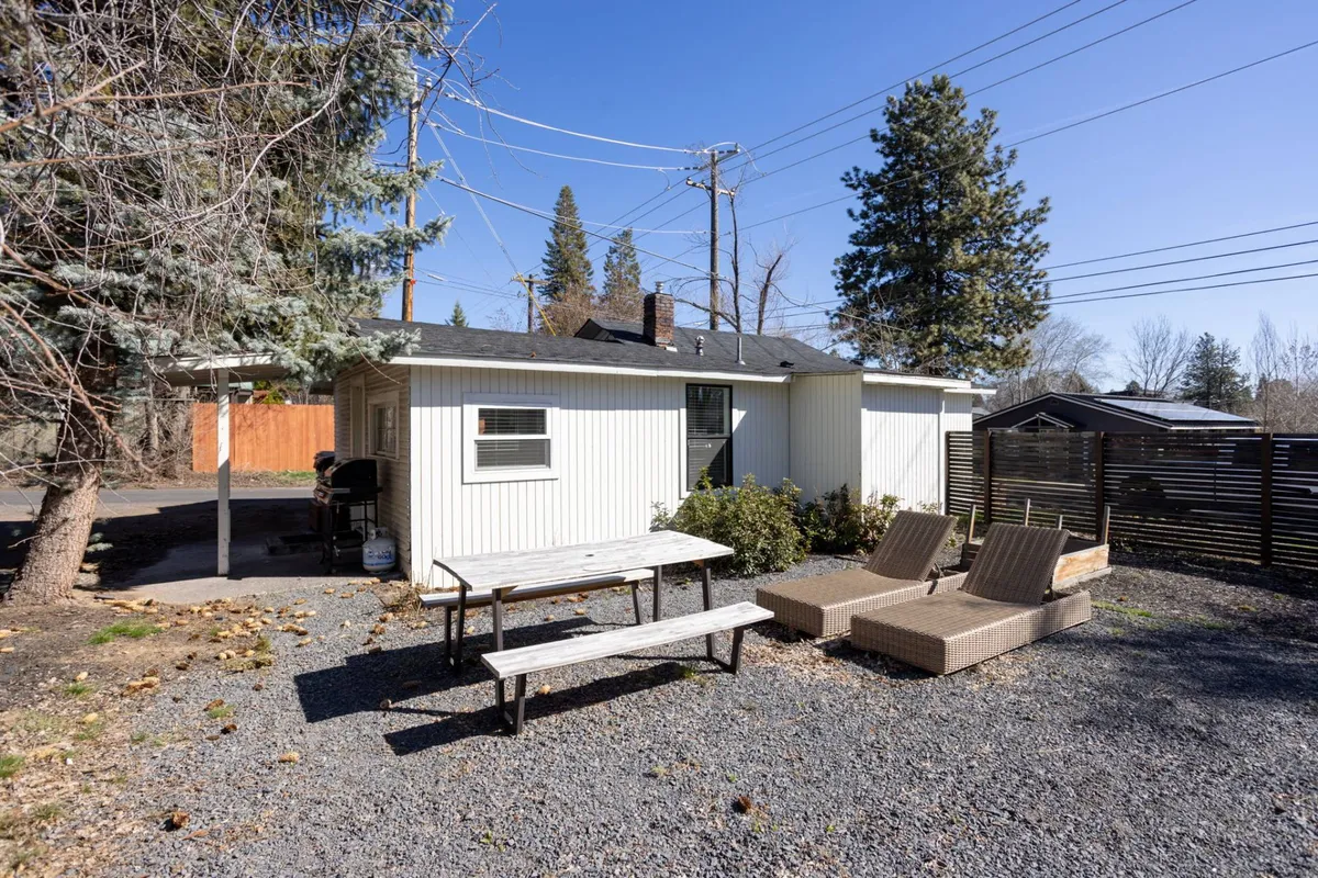 This view presents a fenced backyard with a wooden picnic table, relaxed lounge chairs on a gravel pad, and a border of mature trees. The setting is casual yet private, offering space for dining, sunbathing, or relaxed conversations amid nature.