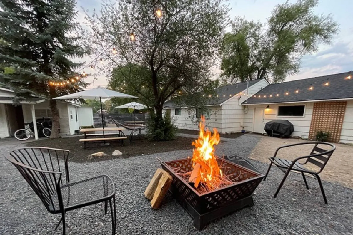 The photo features a glowing fire pit surrounded by inviting chairs and picnic tables, all set beneath twinkling string lights and enveloped by leafy trees. The intimate setup exudes warmth and the perfect ambiance for evening gatherings or storytelling under the stars.